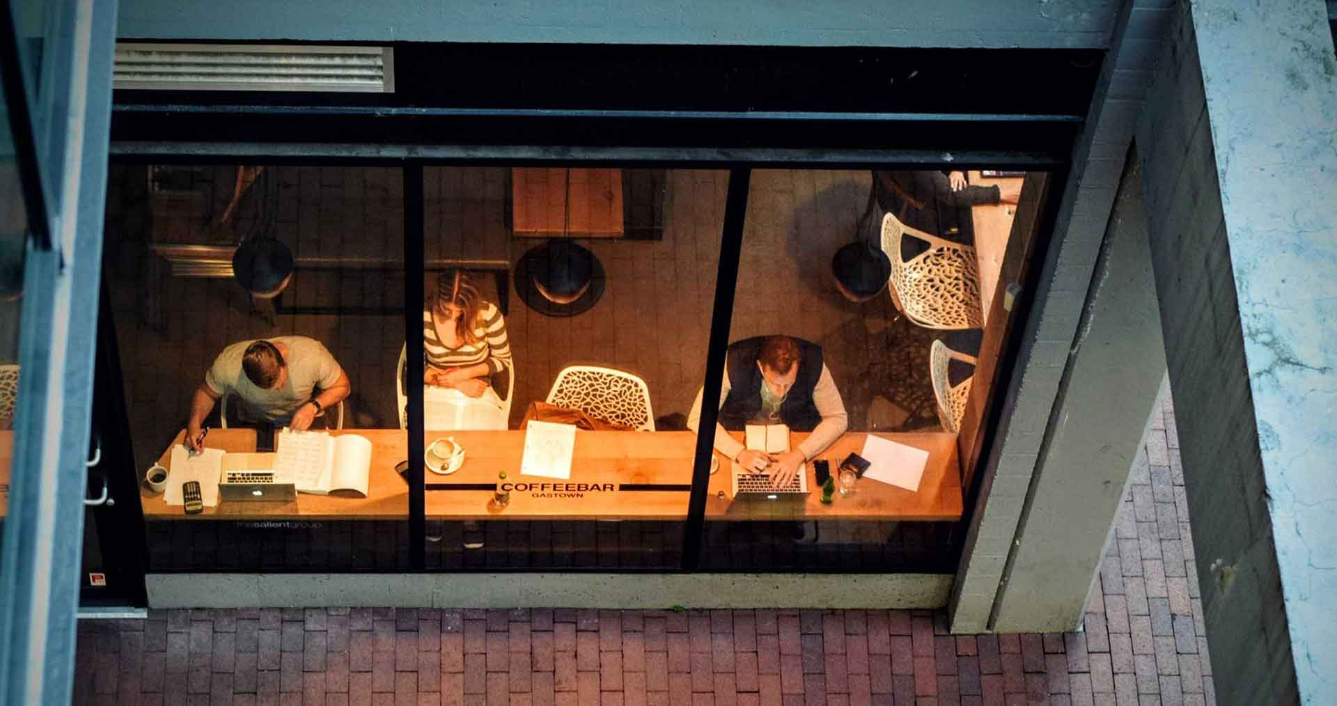 Kahvilassa kaksi miestä kirjoittaa kannettavalla ja nainen lukee. Kuva on kuvattu jyrkästä yläviistosta ison ikkunan läpi.